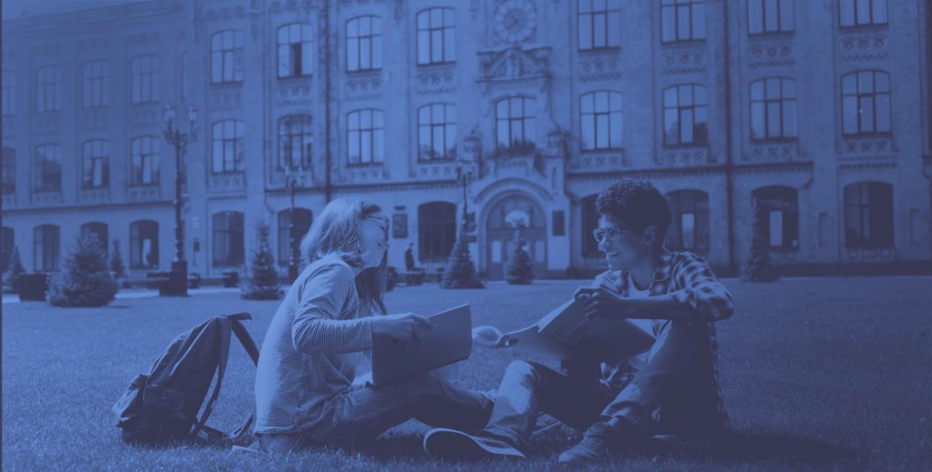 two students sitting on a college campus lawn with backpacks and book smiling